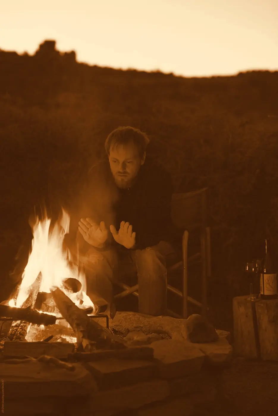 A person warming their hands at a small, contained campfire at dusk