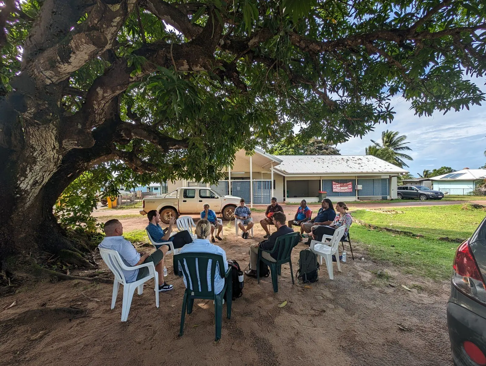 Community members and Emver Partners staff seated in a circle of chairs under a large tree on Badu Island