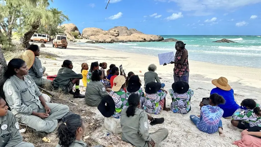 A group of Indigenous rangers and youth seated on a beach in Arnhem Land, listening to a Yolŋu elder presenting a map of Country