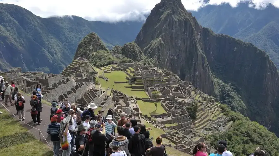 A large crowd of tourists gathered at a viewpoint overlooking Machu Picchu, with the citadel and surrounding mountains in the background