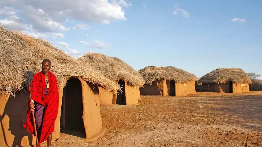 A Maasai man in traditional red shuka standing beside thatched mud-walled homes in a Maasai village at golden hour